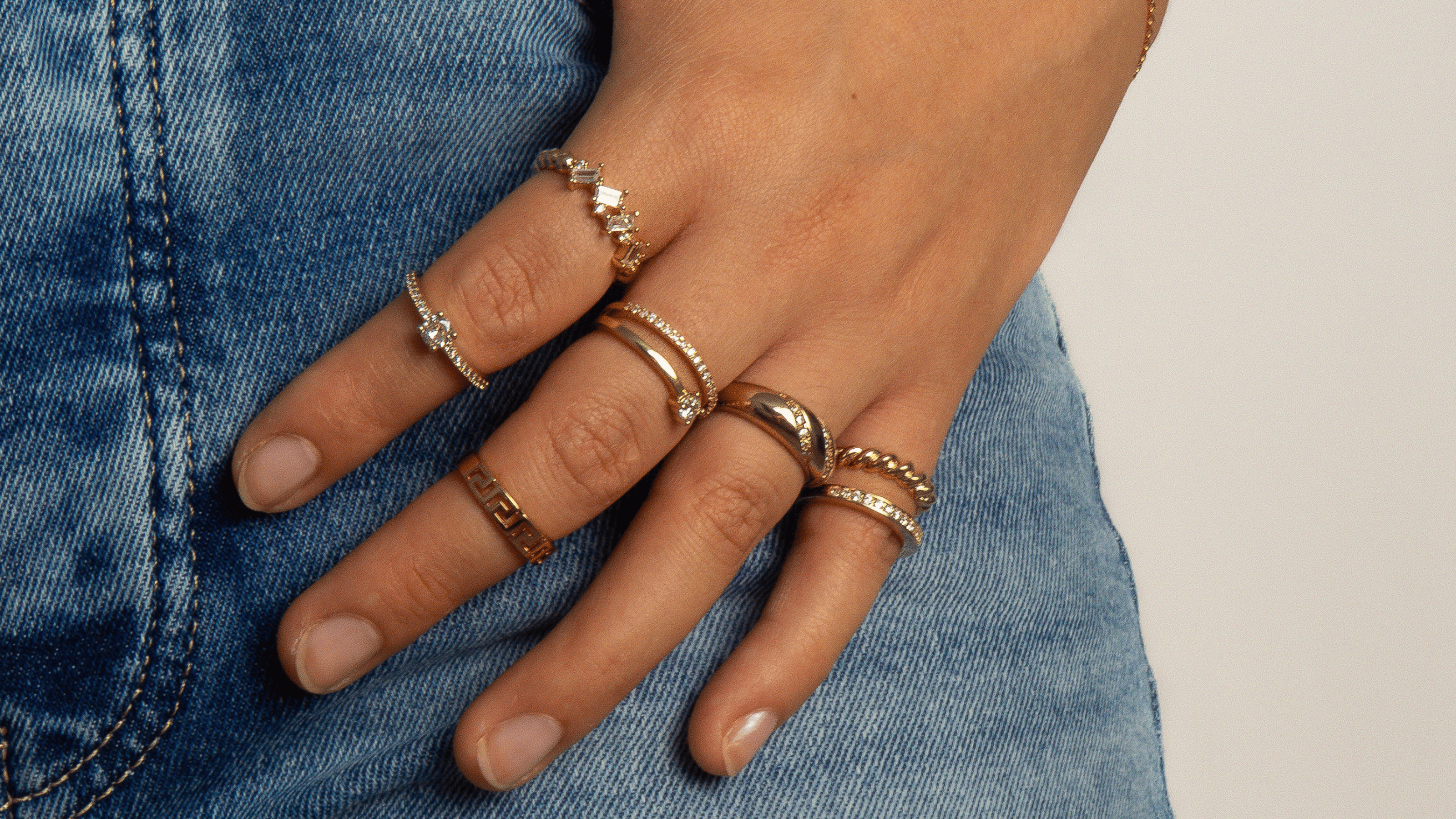 accumulation de bagues en plaqué or et argent 925 portées sur main féminine, avec bracelet fin doré et jean en arrière-plan